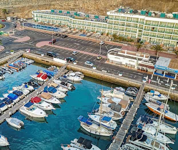 Flygbild av en marina med parkerade båtar framför byggnader och parkering. (AI-genererad text)