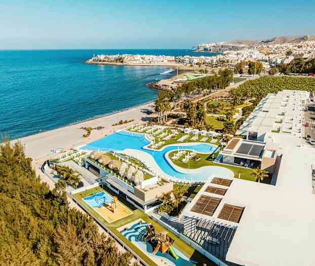 Flygfoto över en kustanläggning med poolområde och byggnader, bredvid en sandstrand och havet. (AI-genererad text)