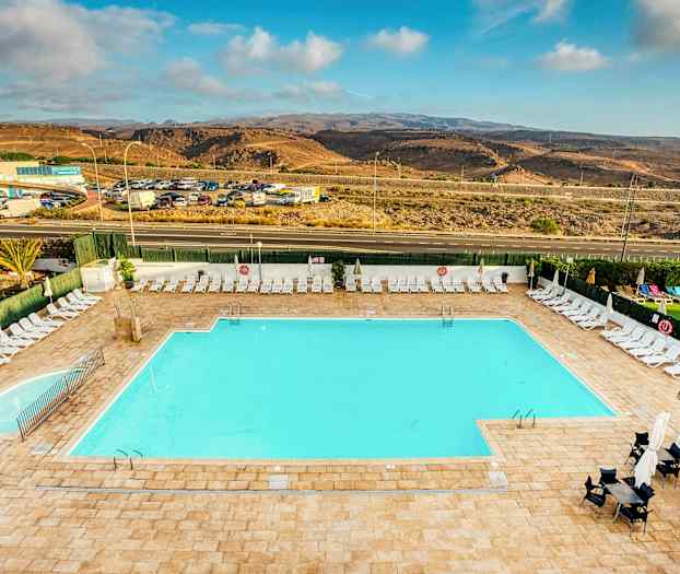 En stor utomhuspool med omgivande solstolar och parasoller. I bakgrunden syns en väg och ett landskap med kullar. (AI-genererad text)