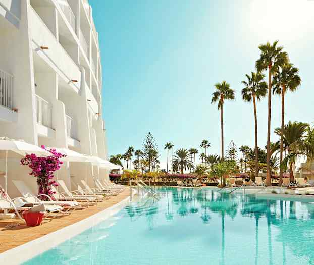 Hotellfasad med balkonger, solstolar vid poolen och palmer i omgivningen. (AI-genererad text)