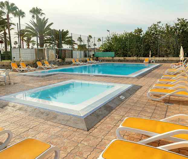 Uteplats med solsängar runt två pooler, omgivna av palmer och staket. (AI-genererad text)