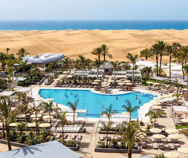 Utsikt över hotellpoolen omgiven av solsängar och parasoller med sanddyner och hav i bakgrunden. (AI-genererad text)