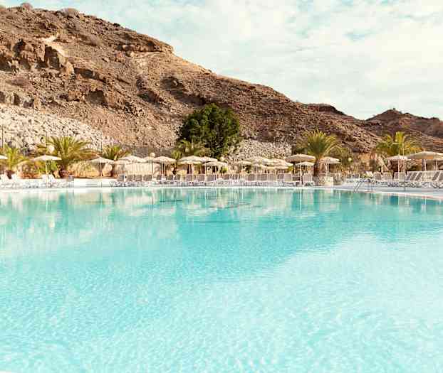Stor pool med solstolar och parasoller vid foten av berg. (AI-genererad text)