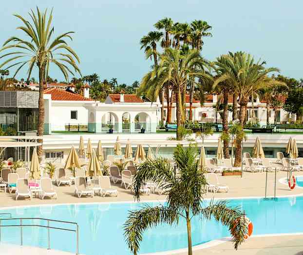 Poolområde vid ett hotell med solsängar och parasoller, omgiven av palmer och byggnader. (AI-genererad text)