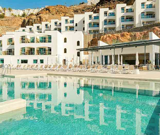 Ett modernt hotell med vit fasad och flera balkonger. Framför hotellet finns en pool och solstolar. (AI-genererad text)