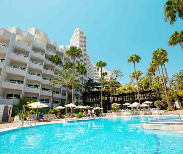 Hotellbyggnad med vit fasad och en stor utomhuspool omgiven av solstolar och parasoller. (AI-genererad text)