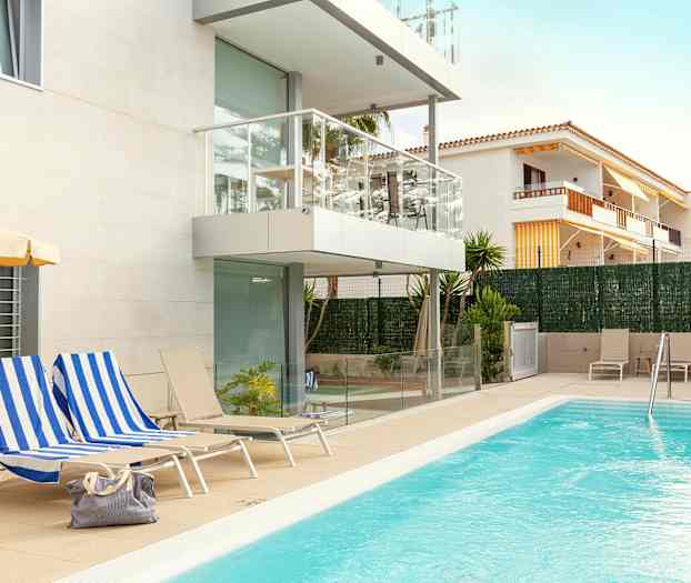 En utomhuspool vid ett modernt lägenhetskomplex med vit fasad och balkonger. Stolar och solstolar finns runt poolområdet och en väska ligger på en av solstolarna. (AI-genererad text)