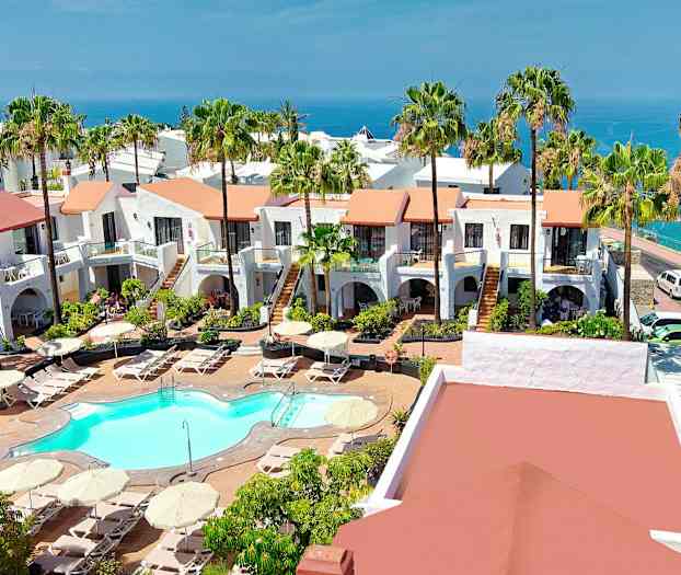 Vy över ett hotellområde med vitkalkade byggnader, en pool och grönskande trädgårdar, havet syns i bakgrunden. (AI-genererad text)