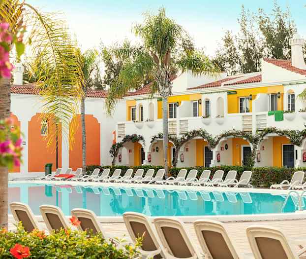 En bild av en pool med solstolar runtom, omgiven av vitmålade och färgglada byggnader med balkonger. Palmträd och blommor syns också. (AI-genererad text)