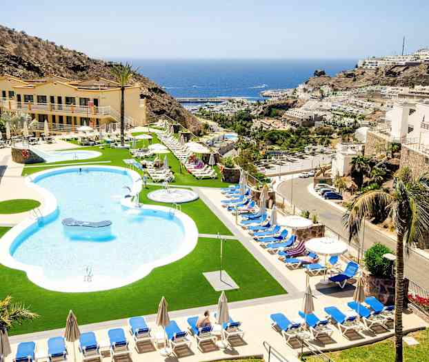 Poolområde med solstolar och parasoller, omgiven av byggnader och berg, med havet i bakgrunden. (AI-genererad text)