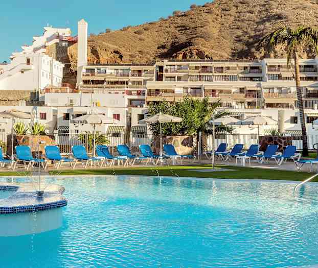 En pool omgiven av solstolar och parasoller framför en byggnad på en sluttning. (AI-genererad text)