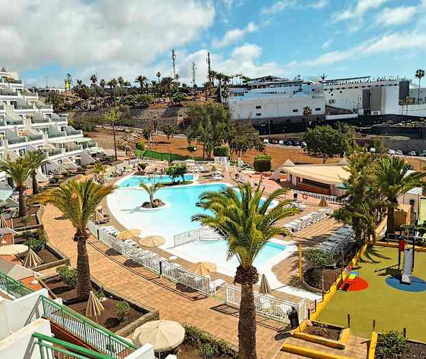 Bild av en semesteranläggning med en pool omgiven av solstolar och parasoller. I förgrunden syns palmer och en lekplats för barn. (AI-genererad text)