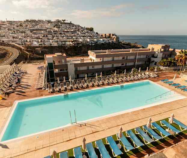 En utomhuspool med solstolar och parasoller vid ett hotellområde, med havsutsikt och bergssluttning med byggnader. (AI-genererad text)