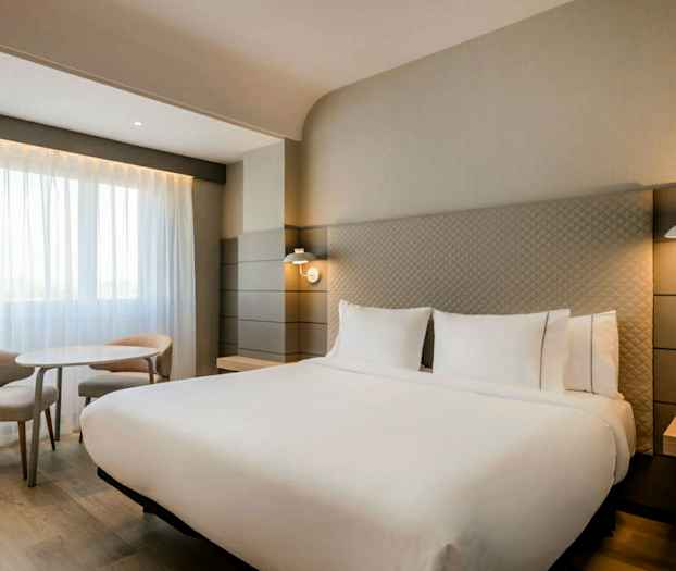 Ett modernt hotellrum med en stor dubbelsäng, två sänglampor, ett bord och två stolar vid ett fönster med ljusa gardiner. (AI-genererad text)