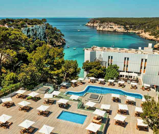 Ett hotellområde med pooler och solstolar ligger vid en vik med klart vatten och klippkust. (AI-genererad text)