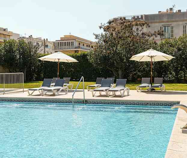 En utomhuspool med blått vatten har solstolar och parasoll på ett kaklat område intill gräsmatta och gröna buskar. (AI-genererad text)