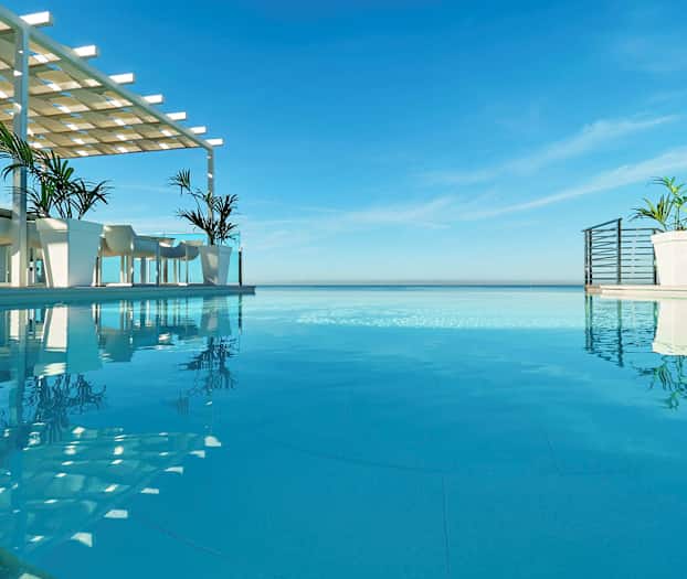 En infinitypool med utsikt över havet, med bord och stolar under en skuggstruktur bredvid poolen. (AI-genererad text)