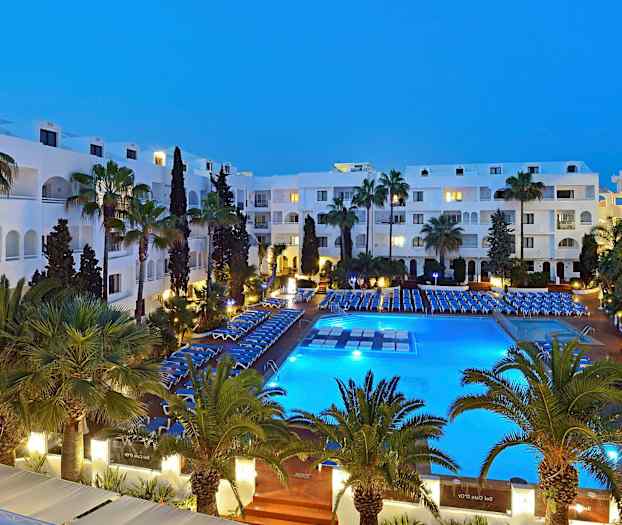 Ett hotellområde på kvällen med en upplyst pool, omgivande stolar och gröna palmer. (AI-genererad text)