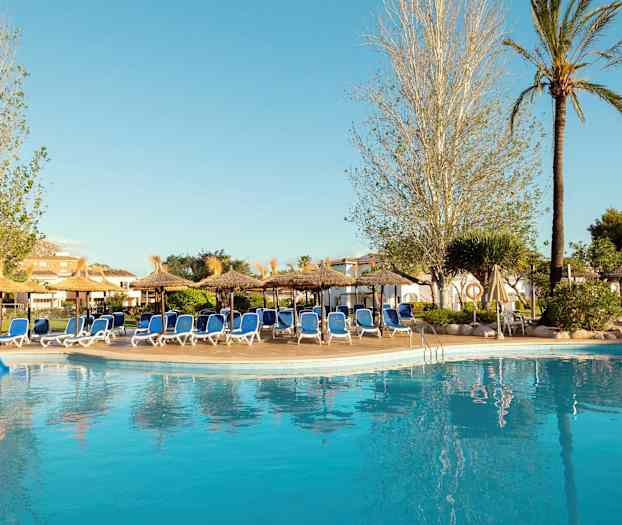 En pool vid ett hotell omgiven av solstolar och parasoller, omgärdad av träd och grönska. (AI-genererad text)