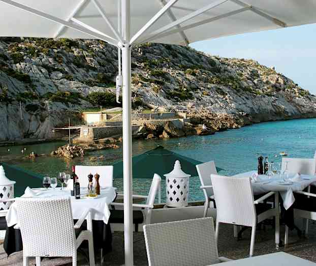 En utomhusrestaurang med dukade bord och vita stolar med utsikt över ett klippigt kustlandskap och havet. (AI-genererad text)