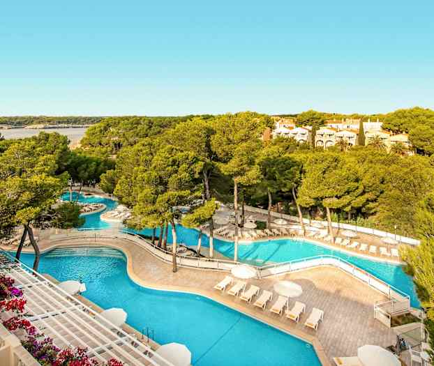 En poolanläggning med solstolar och parasoller omgiven av träd och grönska, med en bukt och bostadshus i bakgrunden. (AI-genererad text)