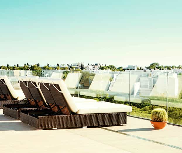 En terrass med solstolar och glasräcken samt utsikt över en modern bebyggelse och grönska. (AI-genererad text)