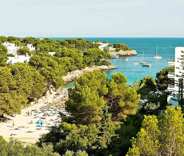 En strand med solsängar och parasoller mellan grönskande träd, och en vik med båtar och klart blått vatten. (AI-genererad text)