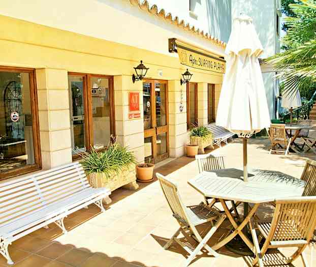 Utomhusterass vid en byggnad med fönster och entré. Trämöbler och en parasoll står på klinkergolvet. (AI-genererad text)