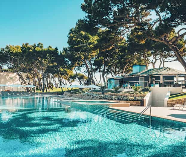 En stor pool omgiven av solsängar och parasoller, med en byggnad och träd i bakgrunden. (AI-genererad text)