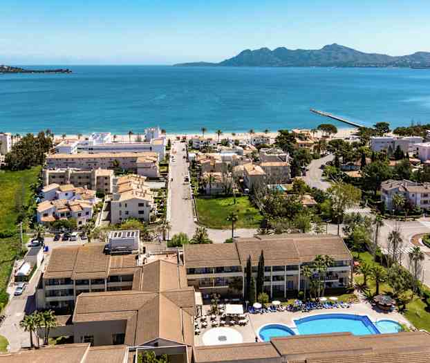 Luftvy över kuststad med hotell, bostadshus och pool i förgrunden, med blått hav och berg i bakgrunden. (AI-genererad text)