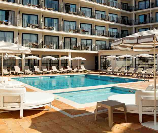 Ett hotellpoolområde med solstolar, parasoller och en pool omgiven av flera våningar med balkonger. (AI-genererad text)