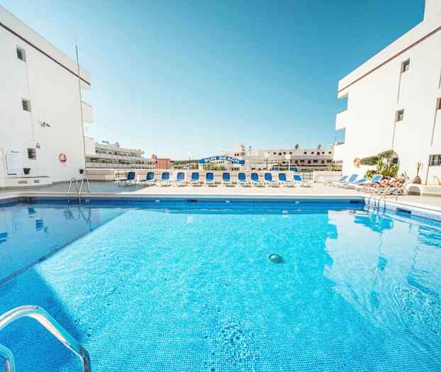 Bild av en swimmingpool omgiven av solstolar och vita byggnader. (AI-genererad text)