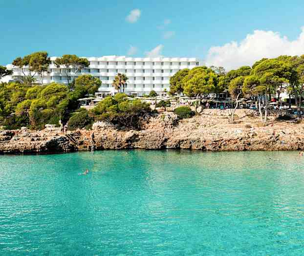 Hotell med vita byggnader bakom en klippig strand med klart blått vatten. (AI-genererad text)