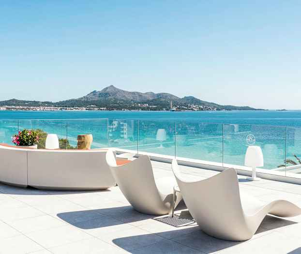 Utsikt från terrass med moderna sittmöbler och glasräcke mot havet och en bergig kustlinje. (AI-genererad text)
