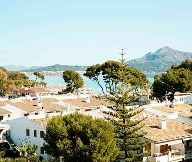 Flera vita hus med tegeltak omgivna av träd, med berg och hav i bakgrunden. (AI-genererad text)