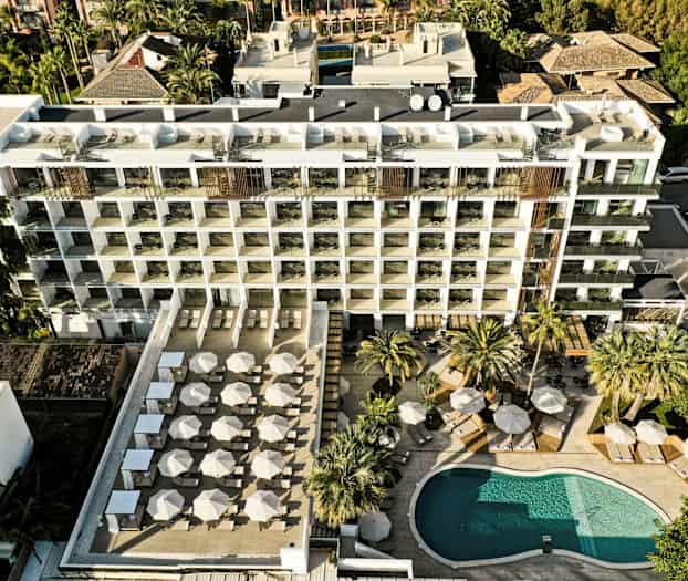 Flygfoto av ett hotell med en pool och en stor terrass med parasoller och solstolar. (AI-genererad text)