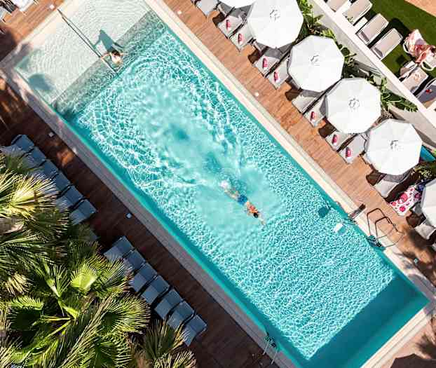 En fågelperspektivbild av en swimmingpool med en simmande person och solstolar med parasoller runtom. (AI-genererad text)