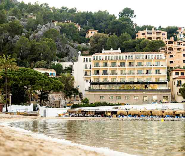 Bild av en strand med klart vatten, en liten restaurang byggnad och flervåningshus omgivna av grönska i bakgrunden. (AI-genererad text)