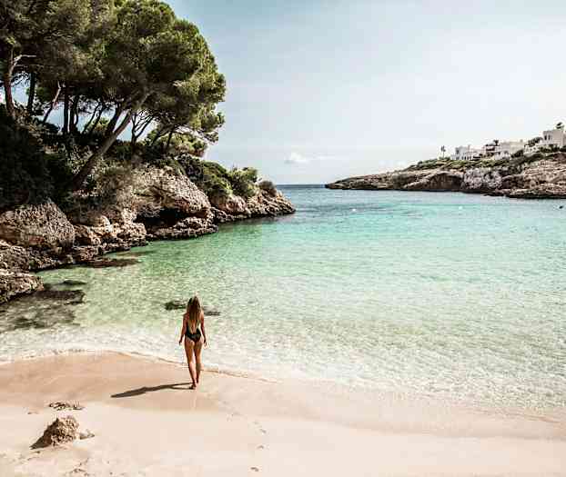 Kvinna står på en sandstrand vid en vik med klart vatten omgivet av klippor och grönska. (AI-genererad text)