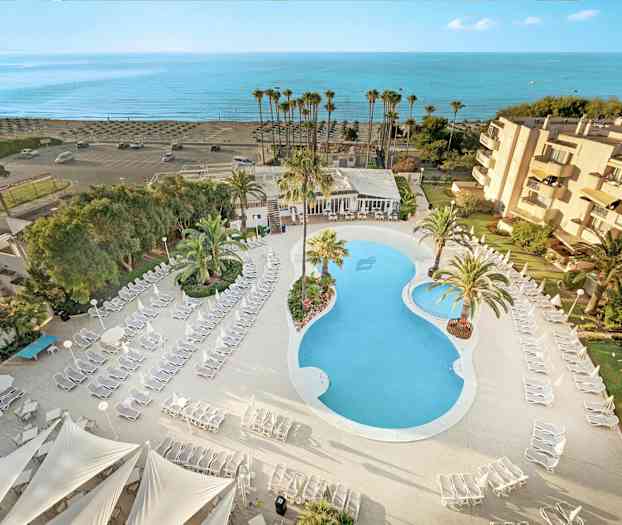 Flygbild över hotellområde med pool, solstolar och palmer, samt närhet till stranden. (AI-genererad text)