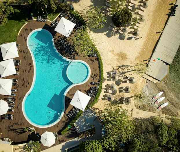 Luftbild över en pool omgiven av solstolar och parasoller, med en angränsande strand och brygga. (AI-genererad text)