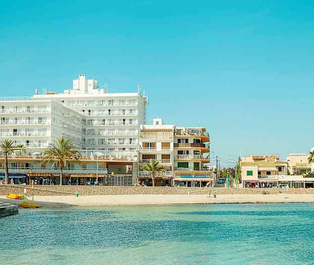 En vy över en strand med klarblått vatten i förgrunden och flera byggnader, inklusive hotell, i bakgrunden. (AI-genererad text)