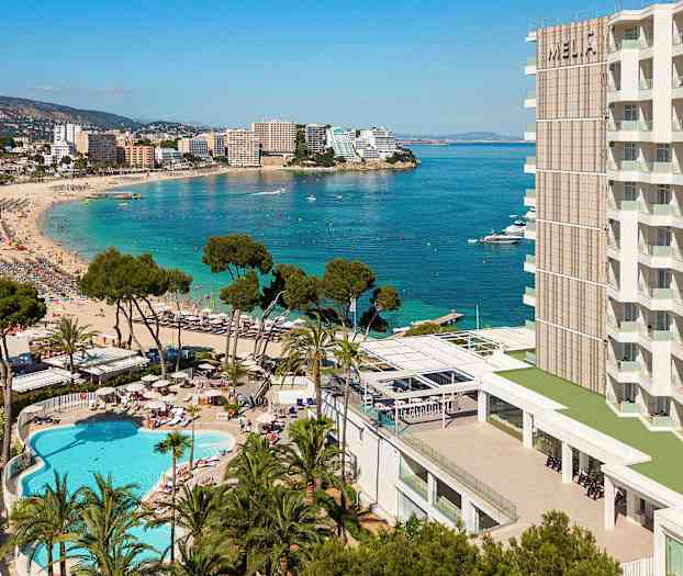 En badstrand med klart blått vatten, omgiven av hotellbyggnader, träd och en pool. (AI-genererad text)