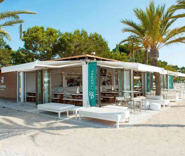 En utomhuslounge och restaurang vid stranden omgiven av palmer. (AI-genererad text)