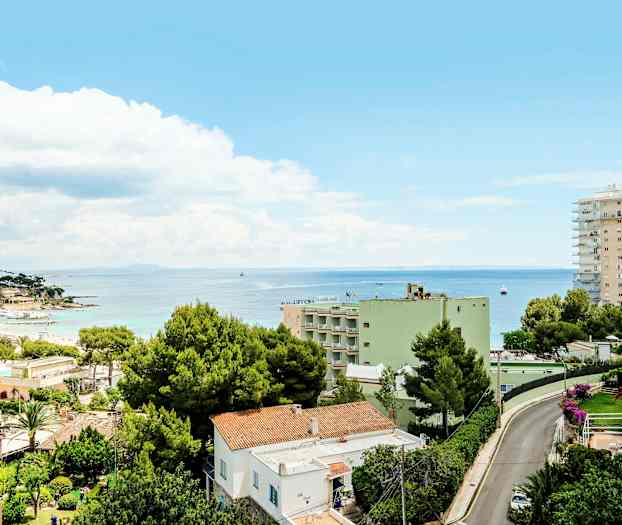 Vy över en stad med bostadshus och gröna träd nära kusten. I bakgrunden syns havet under en blå himmel med moln. (AI-genererad text)