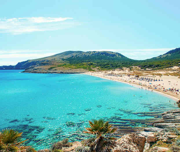 En sandstrand med turkost vatten och grönklädda kullar i bakgrunden. (AI-genererad text)