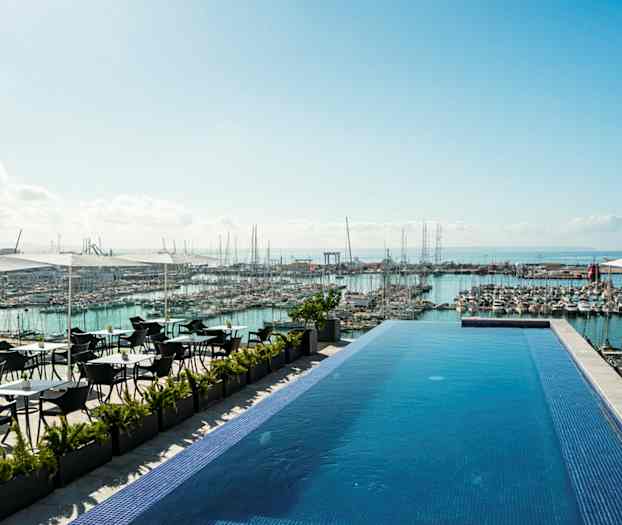 En infinitypool vid en terrass med utsikt över en marina full av båtar och havet i bakgrunden. Det finns sittplatser under parasoller intill poolen. (AI-genererad text)