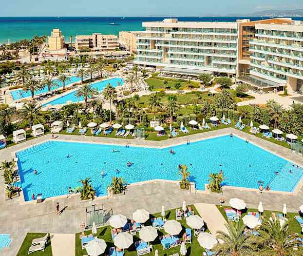 Vy över en semesterresort med pooler, solstolar och omgivande hotellbyggnader, samt havet i bakgrunden. (AI-genererad text)