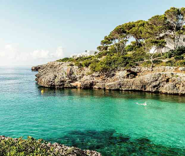 En kustlinje med klippor och klart turkost vatten, ett hus syns bland träden. (AI-genererad text)