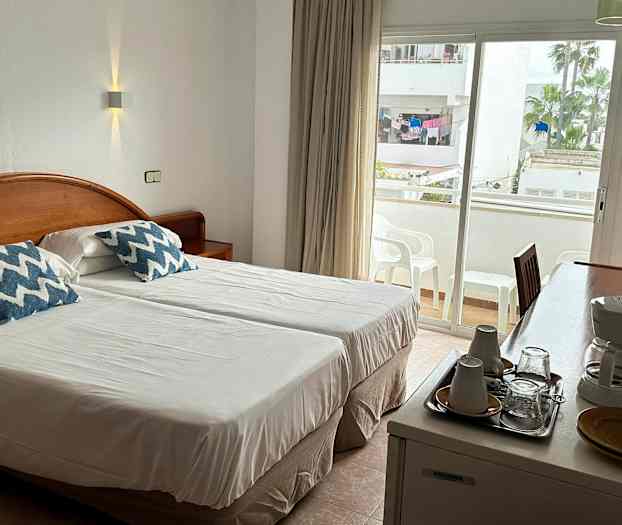 En hotellrum med två enkelsängar, ett skrivbord med kaffekokare och koppar, samt en balkong i bakgrunden. (AI-genererad text)